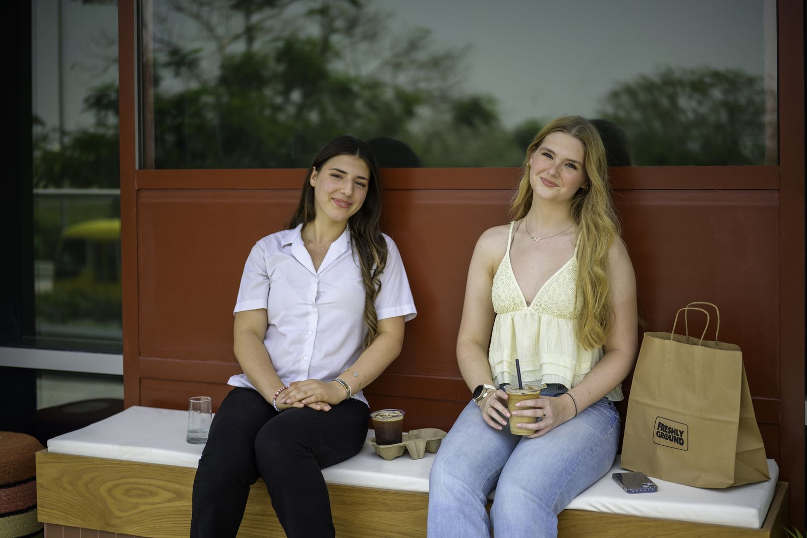 Two guests with iced coffees and a Freshly Ground bag on the terrace bench outside the cafe